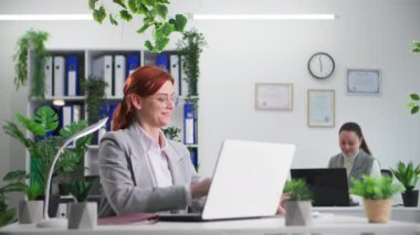 Ofiste çalışırken telefonla konuşan gözlüklü genç bir kadın bilgisayar masasında oturuyor.