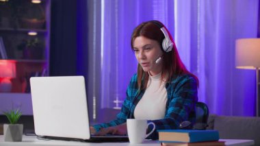 Kulaklıklı, mikrofonlu bir kadın geceleri neon ışıklı odada dizüstü bilgisayarda oyun oynarken...