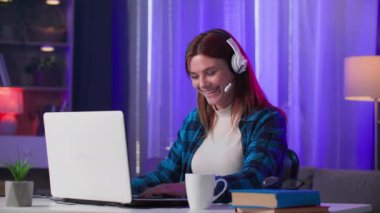 Kulaklıklı bir kadın dizüstü bilgisayarda oyun oynuyor arkadaşlarıyla sohbet ediyor ve evde dinleniyor.