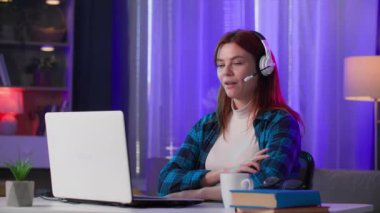 Kulaklıklı genç bir kadının portresi masada oturmuş, neon ışıklarla video oyunları oynuyor ve kameraya bakıyor.