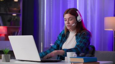 Kulaklıklı neşeli bir kadın, grup sohbetlerinde duygusal olarak iletişim kuruyor. İnternette el sallıyor. Akşamları neon ışıklı odada masada oturuyor.