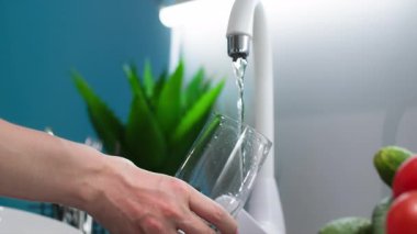 İçme suyu, elinde şeffaf cam olan kadın eli mutfaktaki musluktan soğuk su dolduruyor, yakın plan.