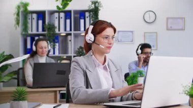Çağrı merkezi, gözlüklü güzel bir kadın video çağrılarına cevap veriyor ve not defterine not alıyor ofisteki meslektaşlarının masasında oturuyor.