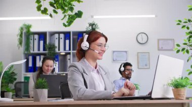 Uluslararası şirket, mikrofon ve kulaklıklı gülümseyen kadın portresi bilgisayarın karşısında oturan kadın ve erkek meslektaşlarının telefonlarına cevap veriyor, kameraya bakıyor.