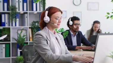 Teknik destek, iş arkadaşlarıyla birlikte bir çağrı merkezinde çalışan ve ofiste otururken mikrofon ve kulaklıkla müşterilerle iletişim kuran gözlüklü genç bir kadın.