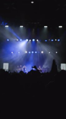 Ön planda el çırpan kadın ellerle alkışlayan bir hayran topluluğu. Bu arada en sevdikleri rock grubu sahnede beyaz ve mavi sahne ışığı altında sahne aldı. Yakın çekim.. 