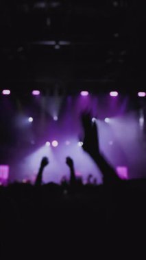 Müzik rengi ışık gösterisi. Orta boy yakın çekim. Sahne ışıklarının karşısında el sallayan hayranların karanlık silueti. Önplanda genç kadınlar yükselen eller ve güçlü duygularla dans ediyorlar.