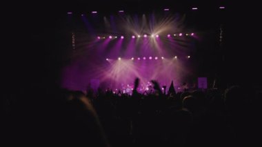 Müzik rengi ışık gösterisi. Orta boy yakın çekim. Sahne ışıklarının karşısında el sallayan hayranların karanlık silueti. Bazı hayranlar telefonlarını kaldırıyor ve filme çekiyor, diğer hayranlar parmaklarını yapıyor