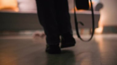 Kot pantolonlu ve botlu bir kadın odanın diğer ucundan kanepeye yürür, çantasını yere bırakır ve yol boyunca botlarını çıkarır. Bacağının gölgesi karanlık gölgesinde.