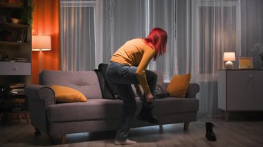 Sarı kazaklı güzel bir kadın aceleyle siyah ceket ve çizme giyerek odanın diğer ucuna atlıyor. Oturma odasında ılık ve loş bir arka ışık var. 