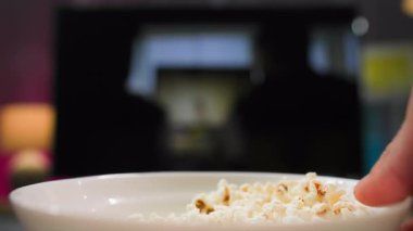 Televizyonun karşısında patlamış mısırı tutan bir adamın aşırı yakın çekimi. Patlamış mısır yerken televizyon izliyor ve elleriyle duygusal ve aktif bir şekilde el kol hareketi yapıyor.
