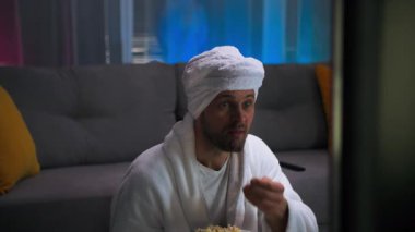 Beyaz bornoz ve havlu giymiş yakışıklı bir adam gergin bir şekilde patlamış mısır yiyor ve TV ekranında olanlara sert bir tepki veriyor. Ekranda olanlarla tamamen ilgiliydi. Yaşarken televizyon izliyor.