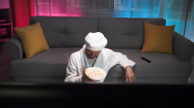 Beyaz bornozlu ve kafasında havluyla çok komik bir adam patlamış mısır yiyor ve televizyon izliyor. Aktif olarak ekranda olanlara el kol hareketi yapıyor ve tepki veriyor. Adam sıcak ve loş bir oturma odasında.