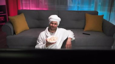 Beyaz önlüklü, patlamış mısırlı, yalnız, orta yaşlı bir adam televizyon izler ve onunla konuşur. Televizyondan adama bak. Pembe ve mavi arka planlı loş ışık oturma odasında.