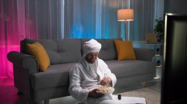 Keyfi yerinde ve gevşemiş bir şekilde uçarken ağzına patlamış mısır atıyor ve televizyon programının tadını çıkarıyor. Oturma odasında loş bir ışık, pembe ve mavi bir arka planla televizyon izliyor.