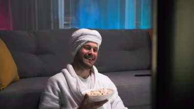 O iyi bir ruh hali içinde, ekranda olanlara sevinçle tepki veriyor, TV programının tadını çıkarıyor. Oturma odasında loş bir ışık, pembe ve mavi bir arka planla televizyon izliyor.