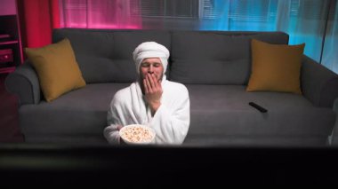 Kafasında havlu olan orta yaşlı bir adam patlamış mısır yiyor, ekranda olanlara aktif olarak tepki veriyor ve gülüyor, televizyondaki kanalları değiştiriyor. Loş ve ılık oturma odası
