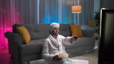 Akşamları tek başına, bir adam yerde oturuyor ve beyaz bir sabahlık ve kafasında bir havluyla sırtını kanepeye yaslıyor. Bir elinde uzaktan kumandayla, diğer elinde patlamış mısırla...