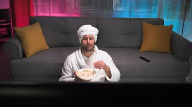 Beyaz önlüklü, yalnız, orta yaşlı bir adam televizyon izlerken ağzına patlamış mısır atıp yakalıyor. Televizyondan adama bak. Pembe ve mavi arka planlı loş ışıklı oturma odası