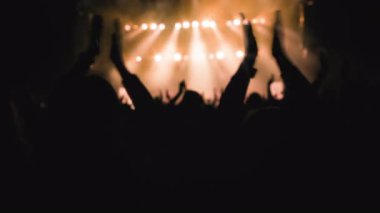 Ön planda el çırpan kadın ellerle alkışlayan bir hayran topluluğu. En sevdikleri rock grubu turuncu flaş ışıkta sahnede çalarken,