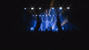 Yakın plan çekim. Ön planda el çırpan kadın ellerle alkışlayan bir hayran topluluğu. En sevdikleri rock grubu sahnede mavi ışıkta çalarken