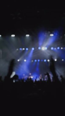 Ön planda el çırpan kadın ellerle alkışlayan bir hayran topluluğu. Bu arada en sevdikleri rock grubu sahnede beyaz ve mavi sahne ışığı altında sahne aldı. Yakın çekim.. 