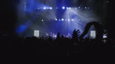 Müzikal renkli ışık gösterisi. Orta boy yakın çekim. Zıplayan kolların koyu silueti sahne ışıklarının altında hareket eden hayranları alkışlıyor. Bir hayran programı telefonundan çekiyor, diğeri de...