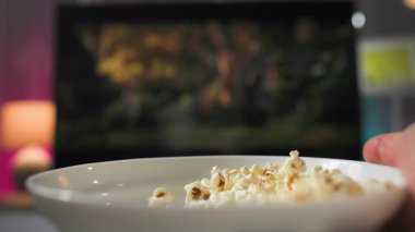 Televizyonun karşısında patlamış mısırı tutan bir adamın aşırı yakın çekimi. O, oturma odasında pembe ve mavi arka planda patlamış mısır yerken televizyon izler..