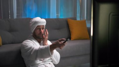 Beyaz önlüklü çekici bir adam, televizyon kumandasını değiştirirken kendine yüz masajı yapıyor. Oturma odasında loş ışıklar ve pembe, mavi arka plan.