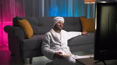 Beyaz bornoz ve havlu giymiş bir adam elinde patlamış mısırla kanepenin yanında oturuyordu. Kaseyi yere koyduktan sonra daha rahat bir şekilde kanepeye uzandı ve kollarını kaldırdı.