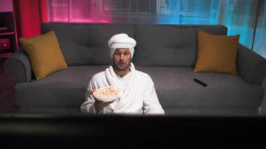 Beyaz önlüklü, yetersiz ve dengesiz orta yaşlı bir adam, kanepenin yanında yerde oturuyor, açık açık konuşmadan televizyonla tartışıyor. Pembe ve loş ışıklı oturma odasında.