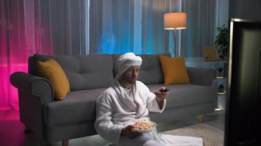 Akşamları tek başına, bir adam yerde oturuyor ve beyaz bir sabahlık ve kafasında bir havluyla sırtını kanepeye yaslıyor. Bir elinde patlamış mısır var ve uzaktan kumandayı elliyor.