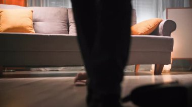 Kot pantolonlu ve siyah çizmeli bir kadın odanın içinde kanepeye doğru yürüyor, çantasını düşürüyor ve yol boyunca botlarını çıkarıyor. Ev ayakkabılarını giyer ve koltuğa oturur.