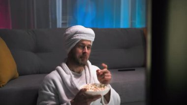 Adamın keyfi yerinde, patlamış mısır yiyor ekranda olanları izliyor, televizyon programının tadını çıkarıyor. O, ekranda olan her şey hakkında yorum yapar. Oturma odasında TV seyrediyor.