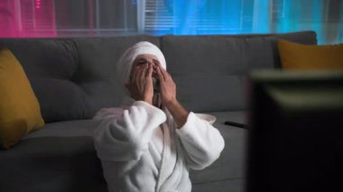 Beyaz önlüklü çekici bir adam güzel yüz maskesi yapıyor, televizyon izlemekten zevk alıyor. Keyfi yerinde ve üşümüş. Loş ışıklar ve pembe ve mavi bir arka planla oturma odasında.