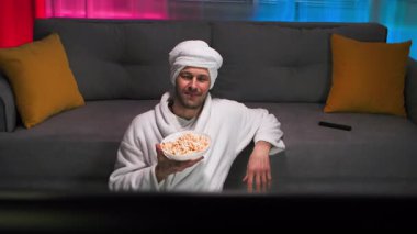 Beyaz önlüklü, patlamış mısırlı, yalnız, orta yaşlı bir adam televizyon izliyor ve uzaktan kumandayla televizyon kanallarını değiştiriyor. Televizyondan adama bak. Pembe ve mavi arka planlı loş ışıklı oturma odasında.