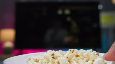 Televizyonun karşısında patlamış mısırı tutan bir adamın aşırı yakın çekimi. O, oturma odasında pembe ve mavi arka planda patlamış mısır yerken televizyon izler..