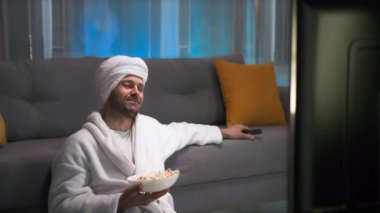 Beyaz önlüklü çekici komik adam kendine yüz masajı yapıyor oturma odasındaki uzaktan kumandayı değiştiriyor loş ışıklar ve pembe, mavi arka plan.