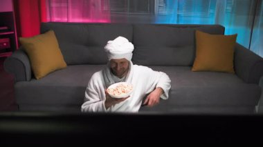 Kafasında havlu olan orta yaşlı bir adam, elinde patlamış mısırla kaseyi tutuyor, ekranda olanlara aktif olarak tepki veriyor ve gülüyor, televizyondaki kanalları değiştiriyor. Loş, ılık, canlı