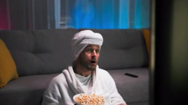 Beyaz bornoz ve havlu giymiş yakışıklı bir adam ağzına patlamış mısır atıyor ve televizyon ekranında olanlara tamamen dalmış durumda. Bir noktadan sonra, neler olup bittiğiyle ilgilenmeye başladı.