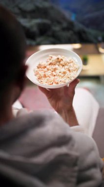 Akşamları elinde patlamış mısırla, yumuşak bir koltukta oturan beyaz ceketli bir adam televizyon izler. Patlamış mısır yer ve sıcak ışıklandırmalı bir oturma odasında televizyon izler.