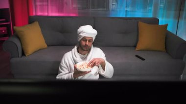 Akşam duştan sonra, beyaz bornozlu komik bir adam kanepenin yanında yere oturur. Patlamış mısır yemekten ve televizyon izlemekten estetik zevk alıyor.