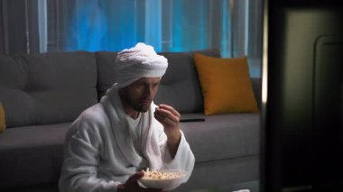 Beyaz önlüklü, elinde patlamış mısırla çekici bir adam TV ekranında olanlara çok şaşırdı ve hayal kırıklığına uğradı. Loş ışıklar ve pembe ve mavi ışıklarla oturma odasında.