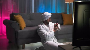 Beyaz bornoz ve havlu giymiş bir adam elinde uzaktan kumandayla koltuğun yanında oturuyor, sonra ekranda olanlara şaşırıyor, uzaktan kumandayı indiriyor ve ne olduğunu anlamaya çalışıyor.