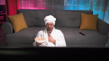 Beyaz bir sabahlık giymiş vahşi, yetersiz ve dengesiz orta yaşlı bir adam kanepenin yanında oturmuş, aktif olarak patlamış mısır yiyor, parlayan gözleriyle TV ekranına bakıyor. Aynı zamanda, bir