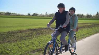 Aktif bir yaşam tarzı sürdüren yaşlı bir çift iki koltuklu bisiklette spor yapıyor. Onlar sıcak bahar güneşli havada güzel bir kırsal alanda bir yol boyunca at sürüyorlar. Bir sürü...