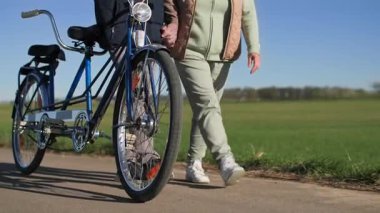 Bisikletli yaşlı bir çift şehir dışında spor gezisi yapıyor. Eşzamanlı hareket ederler ve güneşli bahar havasında yeşil çayırların arka planında el ele tutuşurlar.