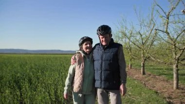 Bisiklet süren yaşlı bir çift, dinlenmek için mola veriyor, kırsal kesimdeki meyve ağaçları boyunca yeşil tarlalarda kol kola yürüyor. Parlak güneşli bir günde ve ılık bahar havasında