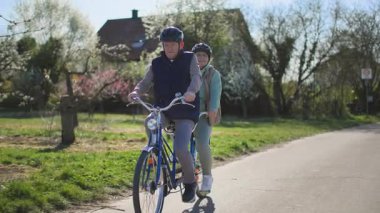Miğfer takan yaşlı bir çift, karı koca, sıcak bahar güneşli havalarda, kırsal bir zeminde iki koltuklu bisiklete biniyor. Bir sürü izlenim ediniyorum.