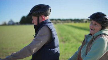 Miğfer takan yaşlı bir çift, karı koca, güneşli havada yeşil tarlaların arka planına karşı iki koltuklu bisiklet sürüyorlar. Bir noktada, adam selamlaşmak için elini sallıyor. Kadın.